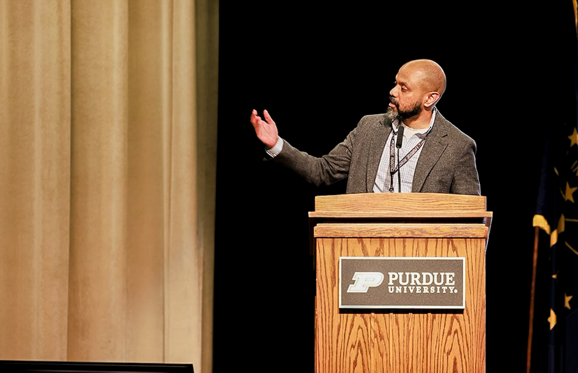 A man speaks at a podium with a Purdue University plaque, gesturing with one hand. The background includes a curtain and part of a flag, conveying a formal event.