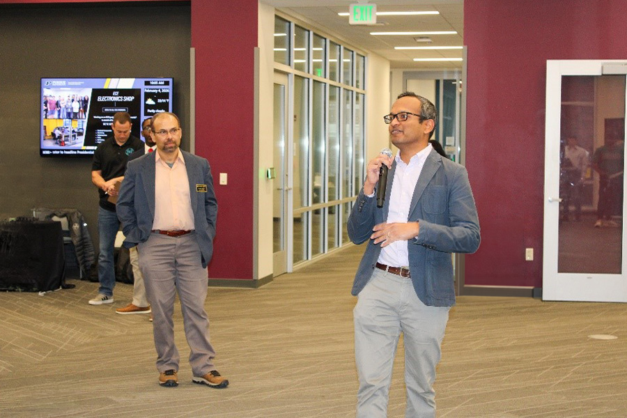 A man in glasses holds a microphone, speaking in a modern office with maroon walls and glass partitions. Another person listens nearby, creating a professional atmosphere.