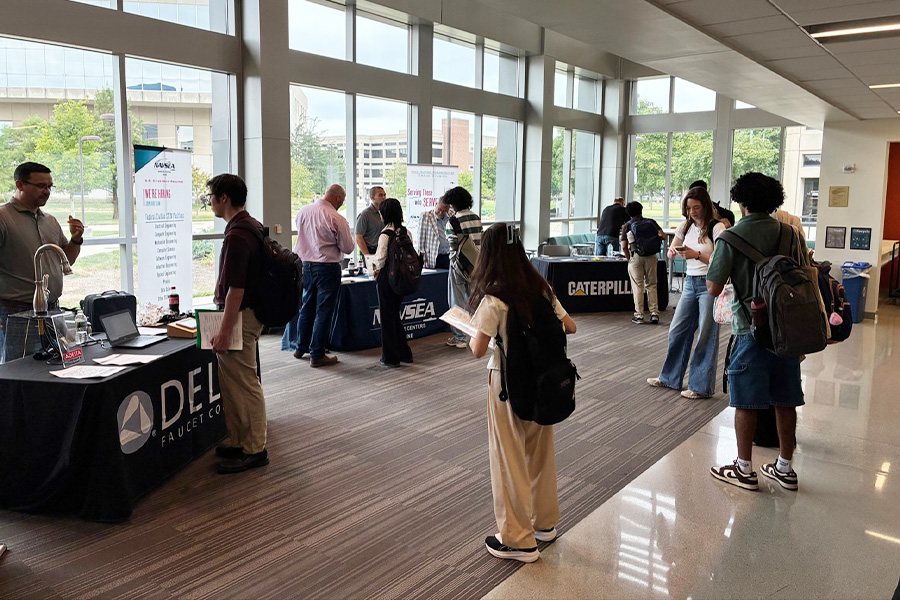 Purdue students speak with company representatives at employer booths during an ECE career fair in a bright campus hallway.