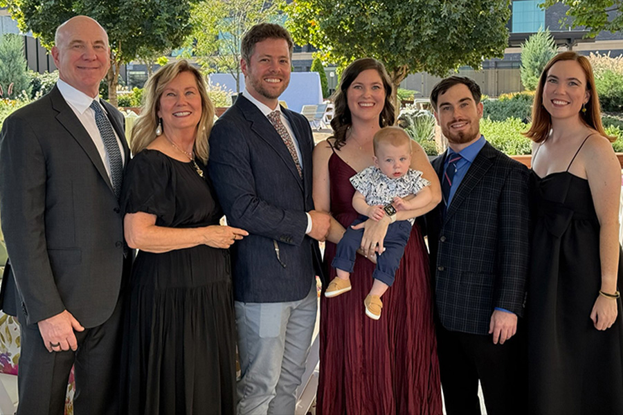 Six well-dressed adults and a baby pose together outdoors at a formal event, standing in front of trees and a modern building. One woman holds the baby while the others stand close together, smiling at the camera.