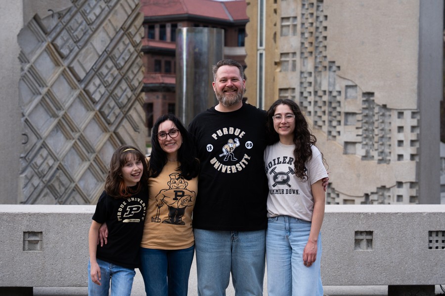 A smiling family of four poses, wearing Purdue University shirts. They stand in front of architectural structures, conveying school pride and togetherness.