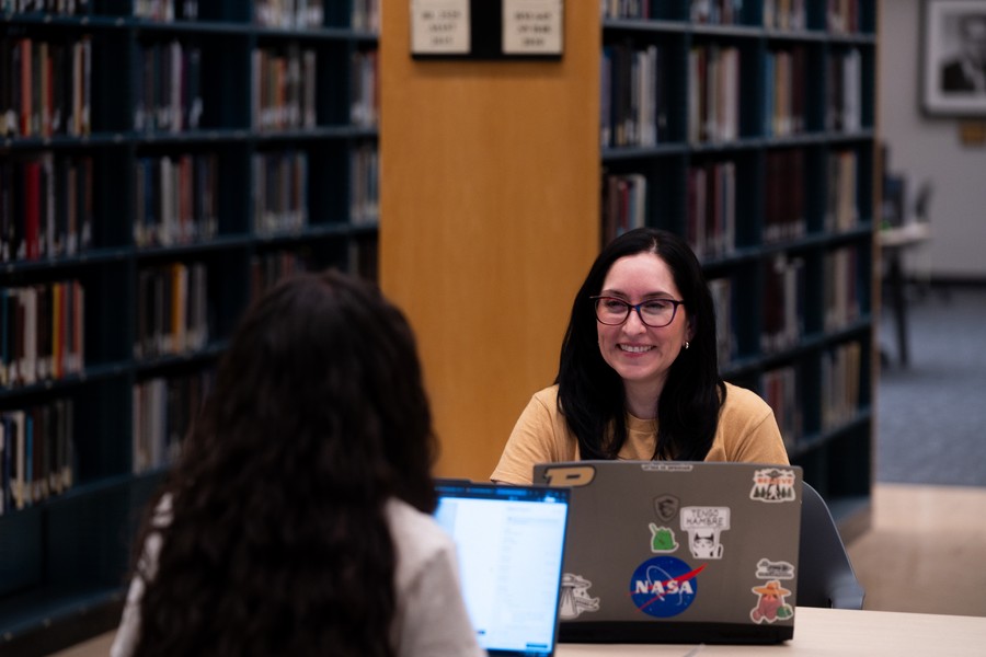 Two people sit across from each other at a library table, both using laptops. One smiles warmly. Shelves of books line the background.