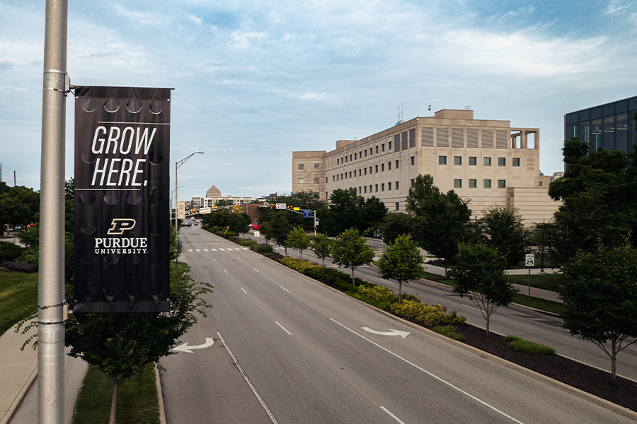 Alt text: Purdue University street banner reading Grow Here hangs from a light pole along a tree-lined city street, with campus buildings visible in the background.