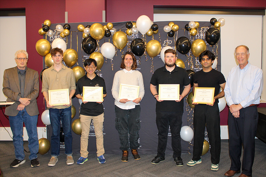 A group of six people pose in front of a Purdue University backdrop with black, gold, and white balloons. Four individuals in the center proudly hold certificates.