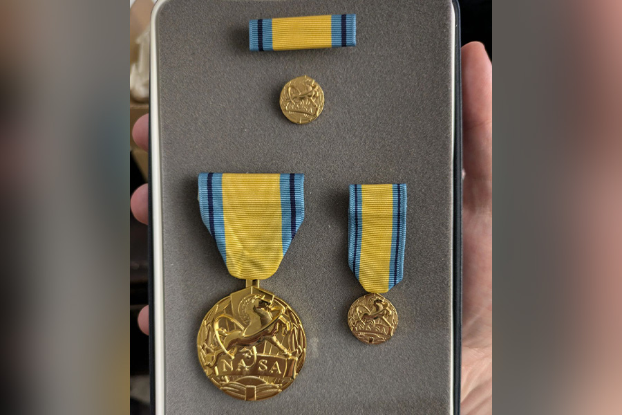 A hand holds a case displaying a gold NASA medal with a blue and yellow ribbon, accompanied by two smaller matching medals and a service ribbon.