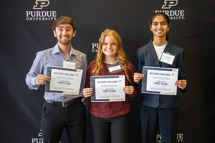 Three Purdue University students smile while holding The Spark Challenge first place certificates in front of a black Purdue University backdrop, celebrating an engineering competition award.