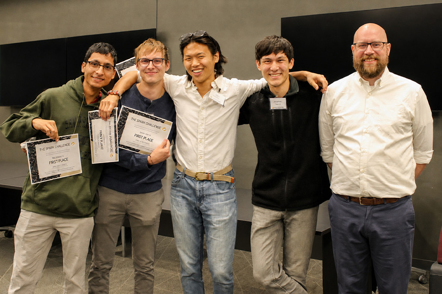 Five people pose together at a Purdue Spark Challenge event, with two participants holding first place certificates and the group smiling in a classroom presentation space.