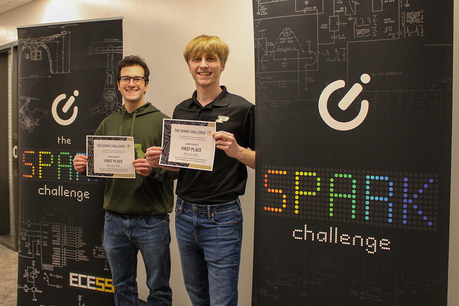 Two Purdue students hold Spark Challenge first place certificates beside ECE-themed competition banners with colorful SPARK challenge text and circuit diagram graphics.