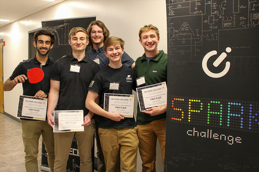 Five Purdue students smile with Spark Challenge first place certificates beside black ECE competition banners, colorful SPARK challenge text, circuit graphics, and a red ping pong paddle.