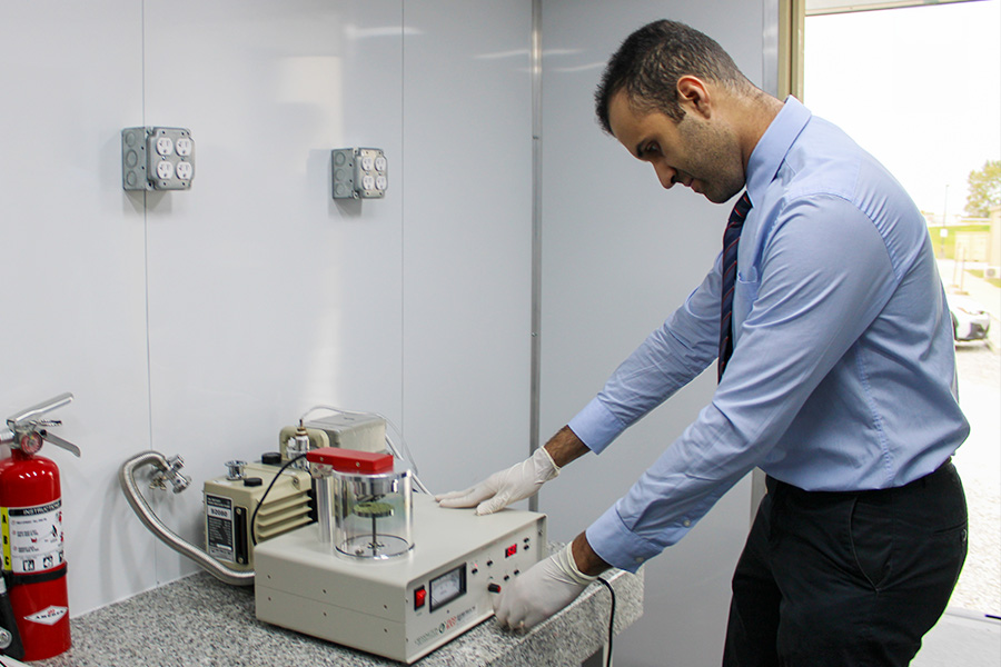 A researcher wearing gloves adjusts laboratory equipment on a countertop while standing beside a vacuum system in a bright lab.