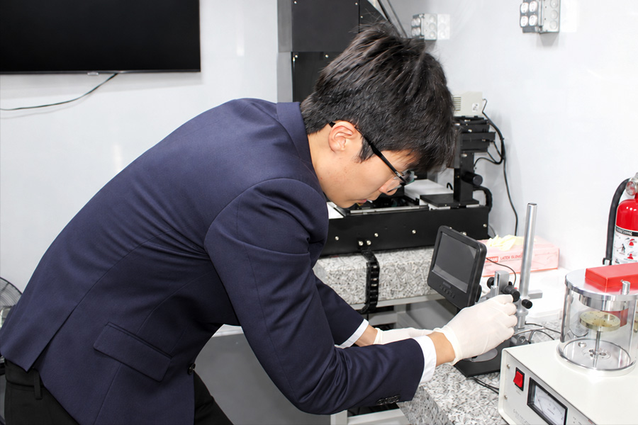 A researcher wearing a navy suit and white gloves works with precision equipment in a laboratory. A black wall-mounted monitor and additional lab instruments are visible in the background.