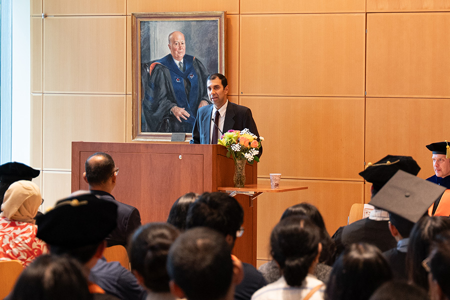 Man giving a formal speech at a wooden podium during a university graduation ceremony, with audience members in caps and gowns, faculty seated on stage, flowers, and a portrait on a wood-paneled wall.