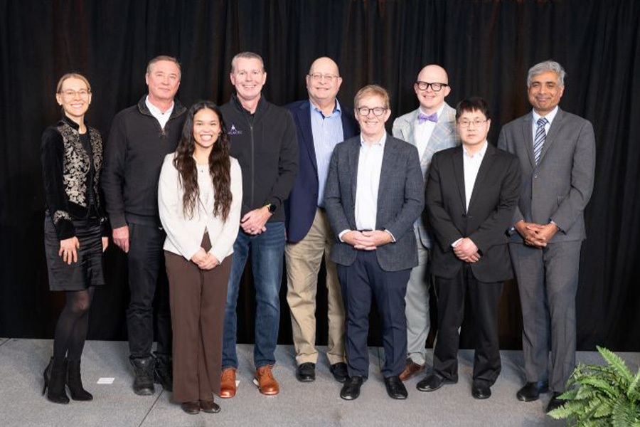 A group of nine people stands side by side, smiling at the camera. They're dressed in business and casual attire, against a black curtain backdrop.
