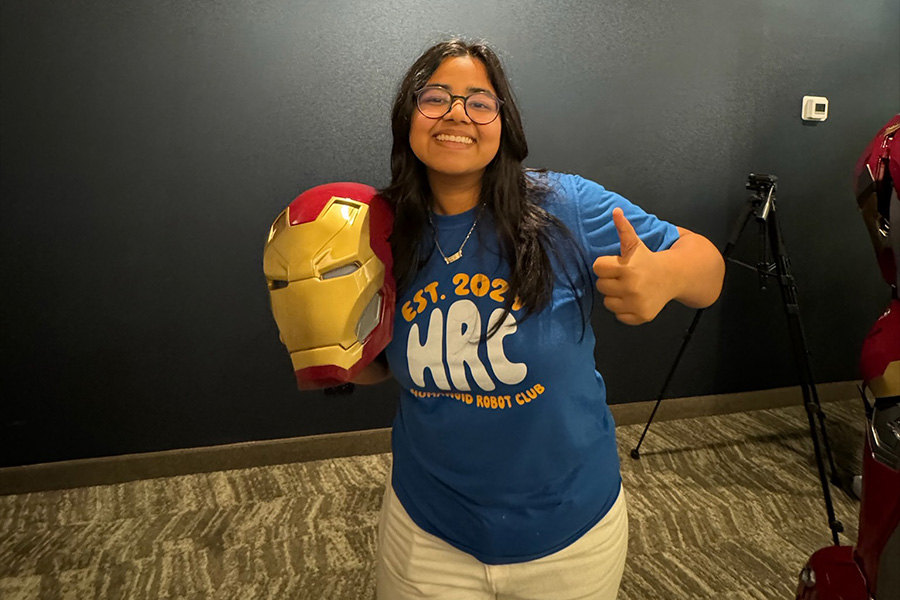 A smiling person in a blue t-shirt holds an Iron Man helmet in one hand and gives a thumbs-up with the other. The setting is indoors with a tripod in the background.