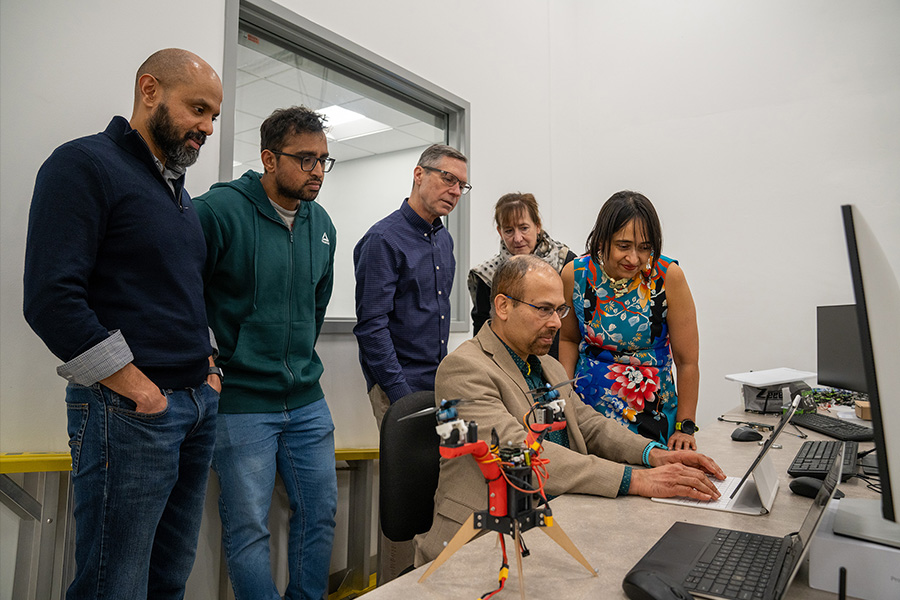 A group of five people gathers around a table, intently watching a man in a suit using a laptop. Nearby is a small mechanical device. The setting exudes focus and curiosity.