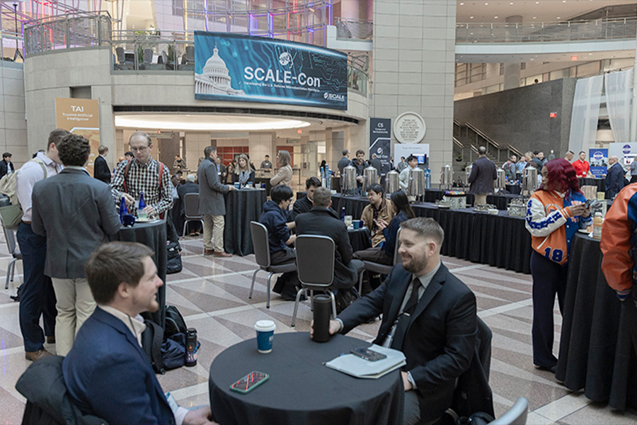 SCALE-Con conference networking event in a modern convention center atrium, with professionals seated at round tables, sponsor booths, coffee stations, and a large digital event banner.