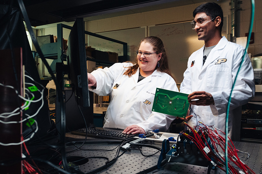Two Purdue students in white lab coats work together in a laboratory. One points at a computer monitor while the other holds a green circuit board, and both are smiling. Lab equipment, wires and testing instruments fill the foreground and surrounding workspace.