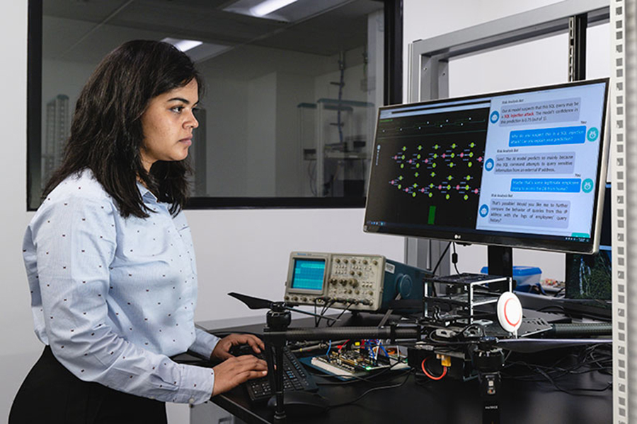 A graduate researcher stands in front of a lab desk. On the desk is a computer monitor and various electrical equipment.