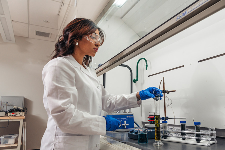 Photo of student in laboratory with blue nitrile gloves on pouring a liquid into a glass container.