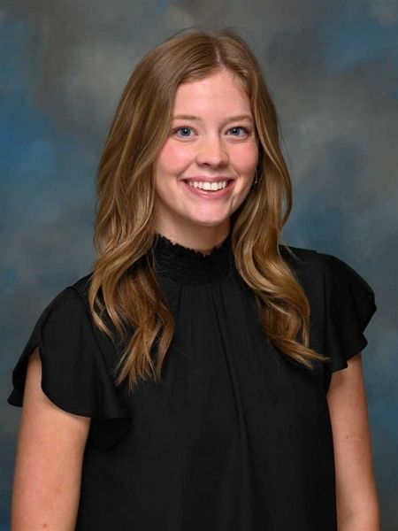 A portrait of Abigail Titus. She is smiling and wearing a black blouse. The background is blue and grey.