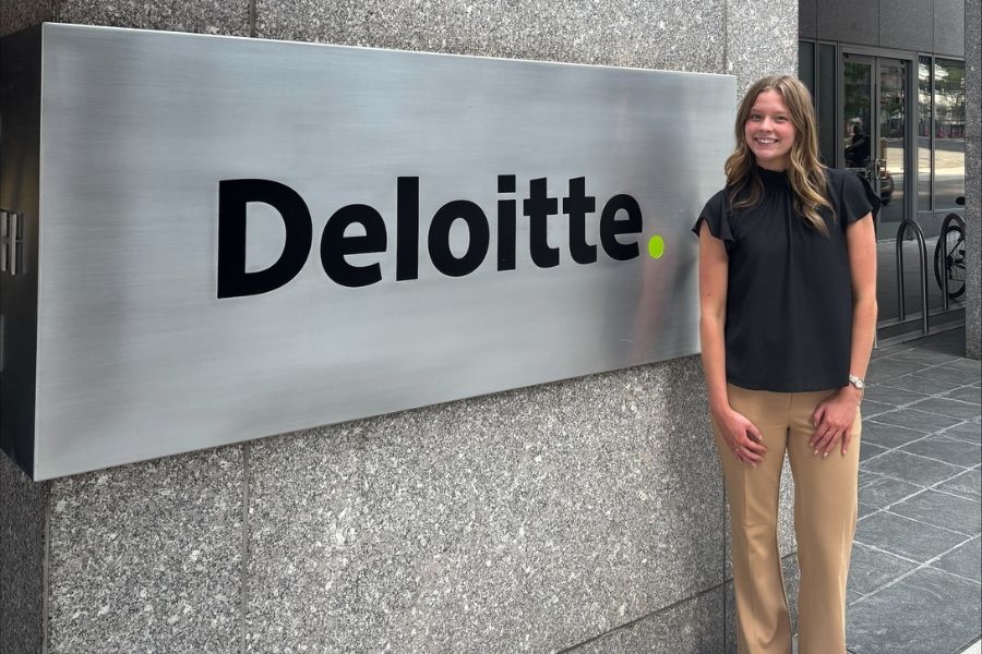 A photo of Abigail Titus standing next to a metal sign with the word "Deloitte." She is wearing a black blouse and tan pants.