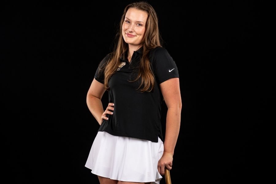 Lane Malek poses with her left hand on the handle of the Purdue hammer and her right hand on her hips. She is wearing a white skirt and black Purdue polo. The background is solid black.