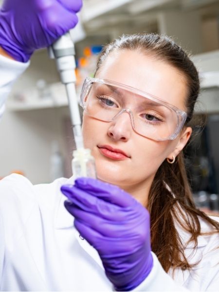 Lana Malek inserts a pipette into a small glass vial. She is wearing clear, protective glasses, purple latex gloves and a white lab coat.