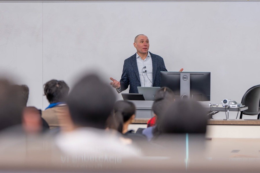 A man in a navy blazer speaks animatedly at a podium in front of a seated audience in a modern classroom, with a Dell monitor and laptop visible at the front of the room.