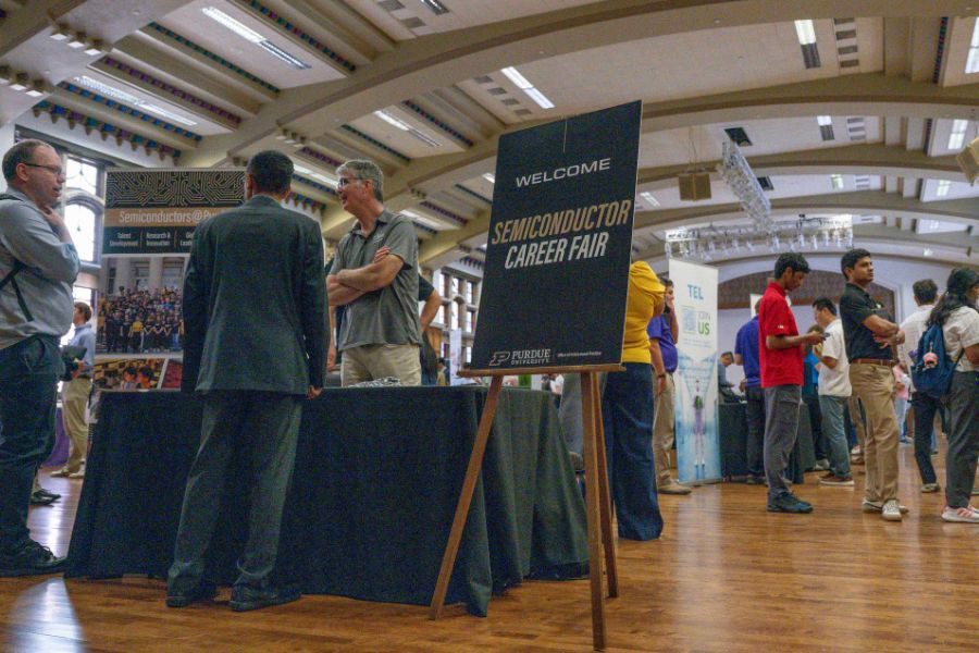Students and industry professionals network at Purdue University's Semiconductor Career Fair, held in a large hall with arched ceilings. A welcome sign on an easel reads "Semiconductor Career Fair" in the foreground, with company booths and attendees visible in the background.