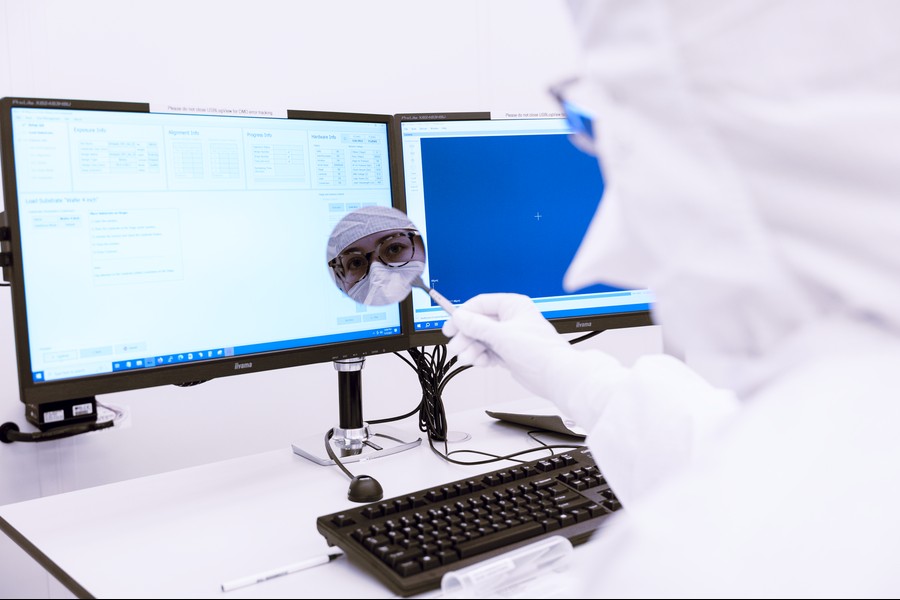 A researcher in full cleanroom protective gear, including a white suit, gloves, face mask, and goggles, uses a tool near a microscope in front of dual monitors displaying semiconductor fabrication software