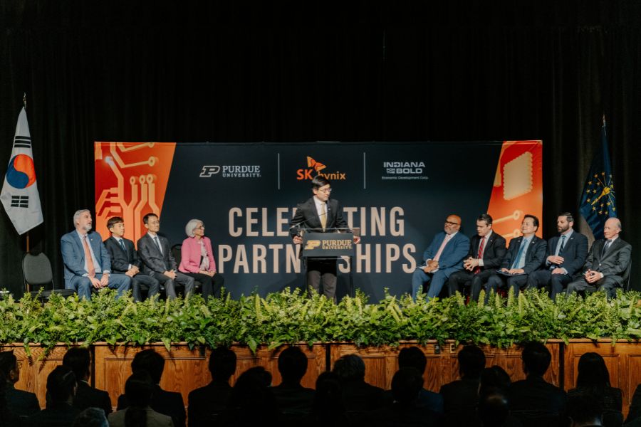 A man in a dark suit and gold tie speaks at a Purdue University podium during a 'Celebrating Partnerships' event, with the SK Hynix logo and an Indiana economic development banner visible in the background.
