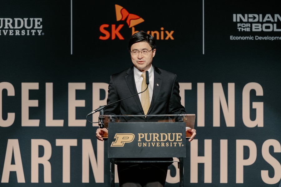 A man in a dark suit and gold tie speaks at a Purdue University podium during a 'Celebrating Partnerships' event, with the SK Hynix logo and an Indiana economic development banner visible in the background.