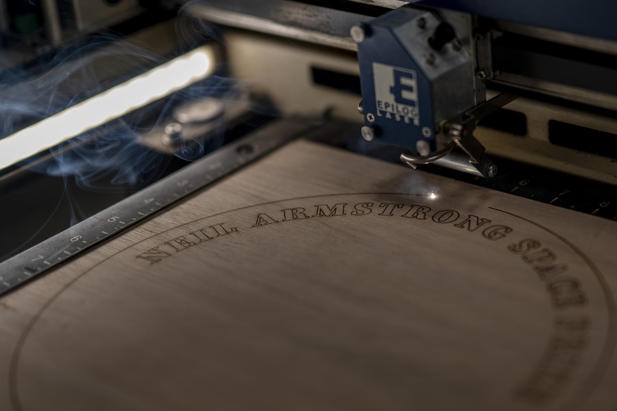 A laser cutter engraves "Neil Armstrong Space Prize" into wood for the medallion's layered shell.