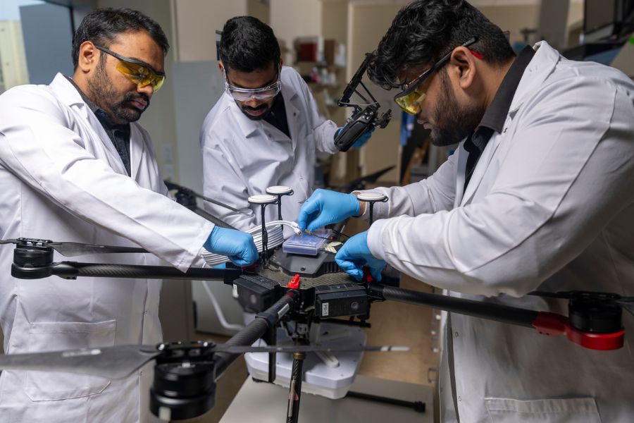Three graduate students in white lab coats work on a large, black drone in Rahim Rahimi's lab.