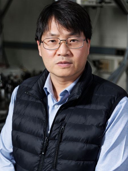 A portrait of Shengwang Du wearing a blue shirt, black vest and glasses.