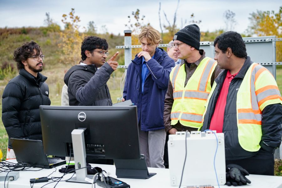A group of students and researchers engage in a discussion while standing in front of an outdoor computer set-up.