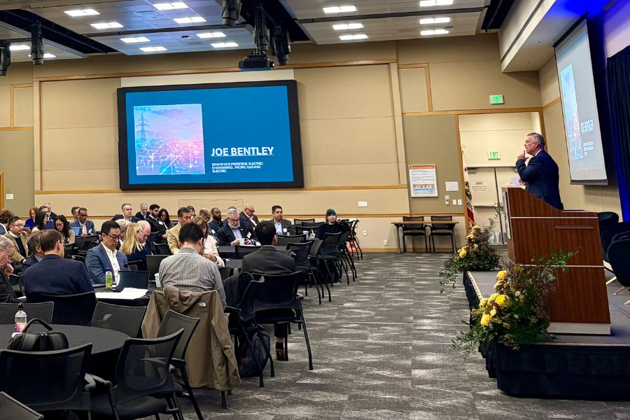 A man in a blue suit stands at an elevated stage in a large conference room in front of dozens of people seated at round tables. 