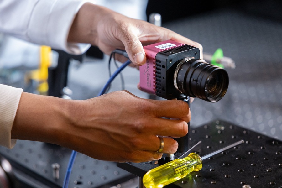 Close-up of two pairs of hands mounting a pink Photon Focus scientific camera with a black telephoto lens onto an optical breadboard. A yellow screwdriver rests on the table nearby. Blue cables and yellow optical mounts are visible in the background.