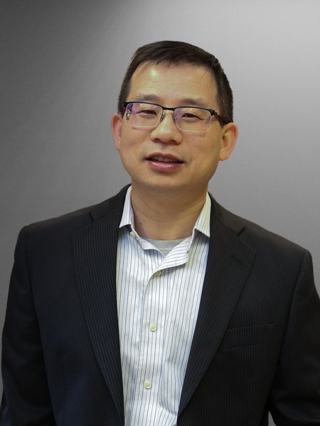Jianguo Mei, Richard and Judith Wien Professor in the James Tarpo Jr. and Margaret Tarpo Department of Chemistry at Purdue University, poses for a professional portrait against a neutral gray background. He is wearing a dark pinstripe blazer over a striped button-down shirt and glasses.