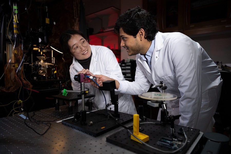 Two researchers in white lab coats lean over an optical table in a dimly lit laboratory. The researcher on the left, wearing glasses, smiles while the researcher on the right adjusts a small pink scientific camera mounted on a post. Optical breadboards, cables, and precision mounts are visible on the table.