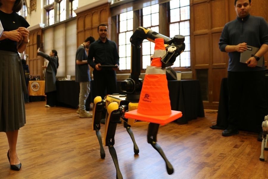 A quadruped robot uses its articulated arm to hold an orange traffic cone while standing on a hardwood floor inside an ornate wood-paneled hall. Several people look on in the background.