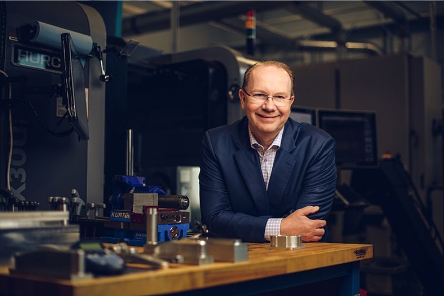  middle-aged man wearing glasses and a navy blue blazer over a plaid shirt leans on a wooden workbench in an industrial machine shop, smiling at the camera. Various machining tools and equipment are visible in the background.