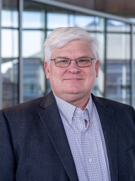 Headshot of a man with white hair and glasses wearing a dark blazer and plaid button-down shirt, posed in front of a glass-windowed building.