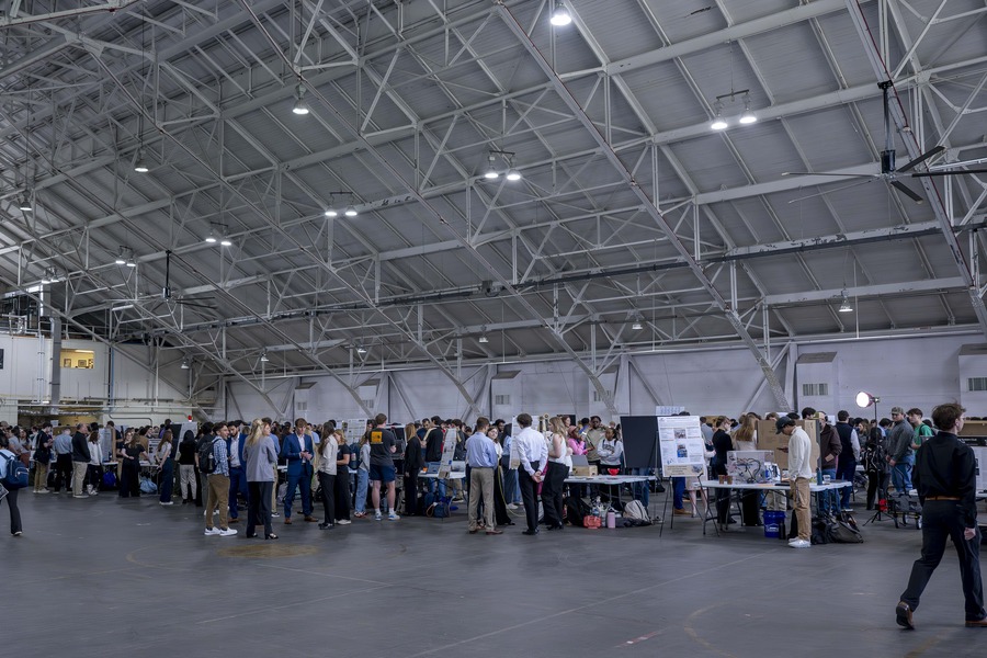 Students and attendees gather around project display tables during an engineering design expo held inside a large industrial hangar. Poster boards, laptops, and physical prototypes are visible at each station as participants present and discuss their work.