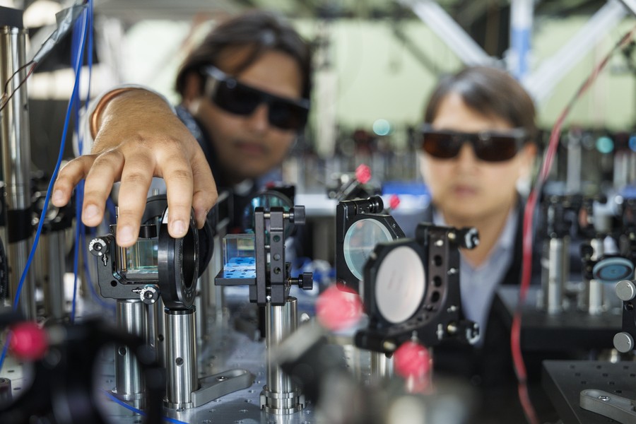 Two researchers wearing laser safety glasses adjust optical components on a photonics lab bench. One reaches toward a beam-splitting optic mounted on a precision post; mirrors, lenses, and fiber optic cables are arranged across the optical breadboard in the foreground.