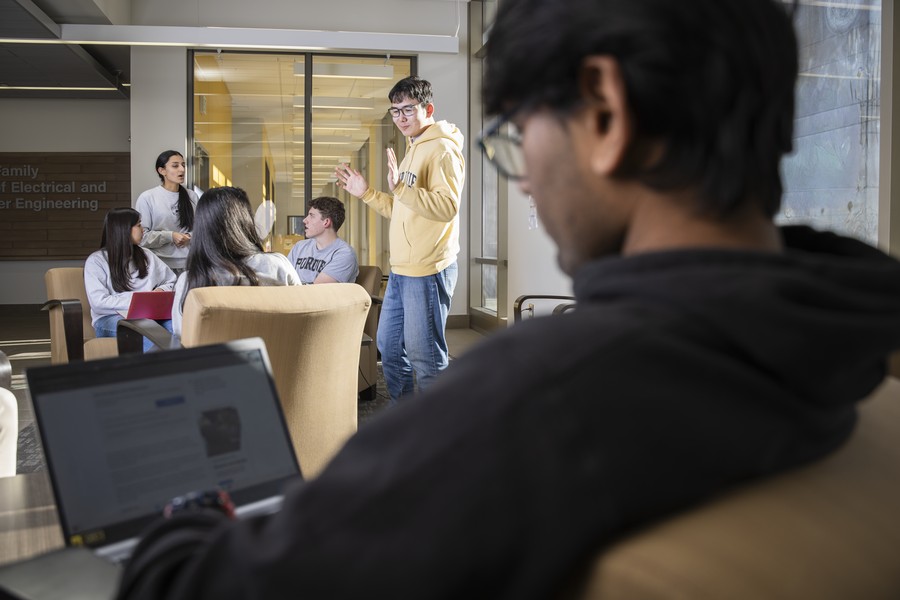 A group of students collaborates in a lounge area. One student in a yellow Purdue hoodie stands and gestures while speaking to several seated peers; a student in the foreground works on a laptop.