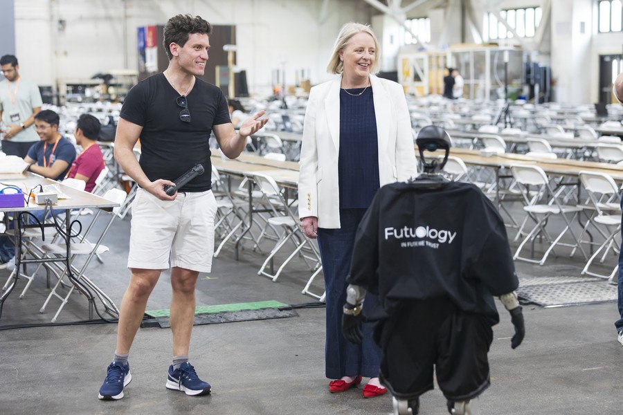 A man gestures while speaking to a woman in a white blazer as a small humanoid robot wearing a "Futology" cape stands between them on the floor of the Purdue University Armory during StarkHacks.