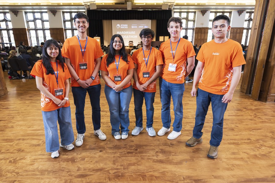 Six smiling students stand together in a large wood-floored hall, all wearing matching orange "builder" t-shirts and lanyards with event badges. In the background, a projection screen reads "Stark Hacks Hardware Hackathon," with rows of seated participants visible. The setting appears to be a university event space with tall windows and ornate wooden architecture.