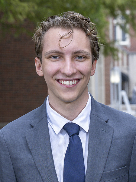 Student with short curly hair and blue eyes, wearing a gray suit and blue tie