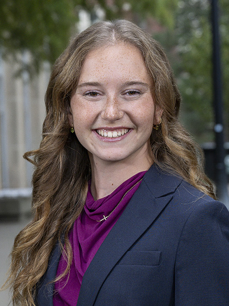 Student long hair, freckles, smiling wearing a navy suit jacket and purple shirt